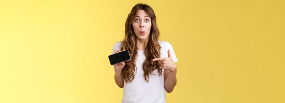 Surprised enthsuastic intrigued cute curly-haired caucasian girl folding lips amused curiously pointing smartphone screen stare camera ambushed astonished describe cool mobile phone app.