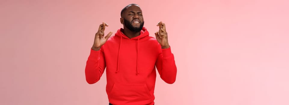 Passionate devoted african-american male fan praying cross fingers good luck asking god win bet biting lip impatiently waiting important results supplicating hopefully, standing pink background.