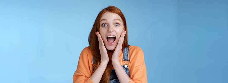 Lifestyle. Excited thrilled young emotional enthusiasitc ginger girl teenage college student yelling amused smiling broadly receive positive good news look surprised camera touch face astonished blue background.