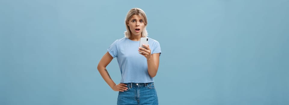 Worried confused young blonde woman in trendy outfit looking concerned and anxious realising bad thing happened receiving shocking news via smartphone opening mouth and staring at camera. Technology concept