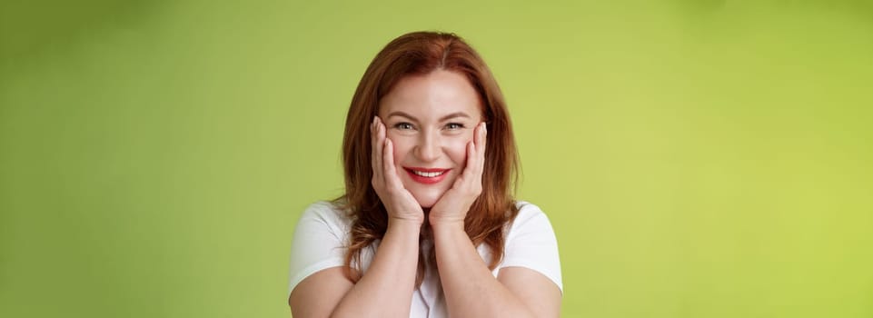 Happy delighted cheerful lucky redhead middle-aged caucasian woman. blushing joyfully receive touching cute gift touch cheeks pleased delighted smiling broadly feel happiness joy green background.