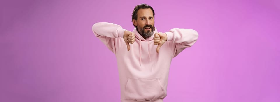Disappointed upset angry caucasian adult bearded male show thumbs down dislking bad unprofessional behaviour frowning demanding refund standing bothered give negative feedback, purple background.