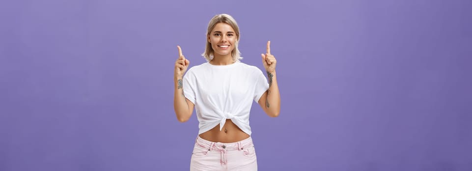 Time look upwards and move forward corporate ladder. Portrait of attractive ambitious and stylish young blonde woman with tattoos and pierced belly pointing up and smiling broadly over purple wall. Copy space