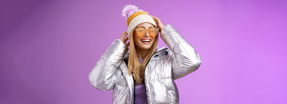 Carefree amused charming blond girlfriend having fun enjoying awesome sunny winter day ski resort vacation wearing sunglasses silver stylish jacket put on hat grinning joyfully, purple background.