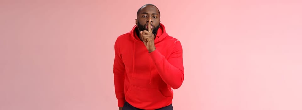 Shhh it taboo. Portrait sensual funny cute african-american bearded man in red hoodie bending camera show shush gesture index finger on lips telling secret asking keep quiet not tell anyone mystery.