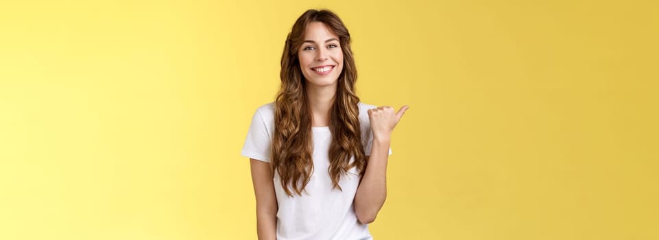 Visit see yourself. Cheerful charismatic good-looking outgoing girl long curly haircut showing place do good hairstyle smiling happily delighted pointing thumb left introduce promo yellow background.