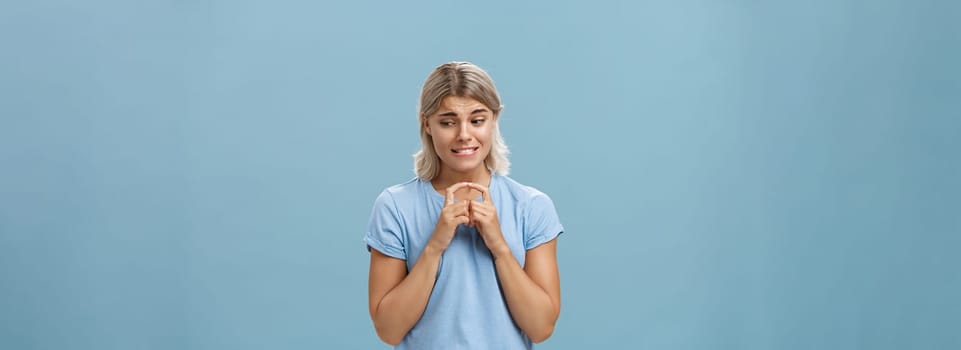 Lifestyle. Studio shot of insecure and timid silly girlfriend with blond hair clenching teeth looking shy down and steepling index fingers unconfident trying ask out boy she likes over blue background.