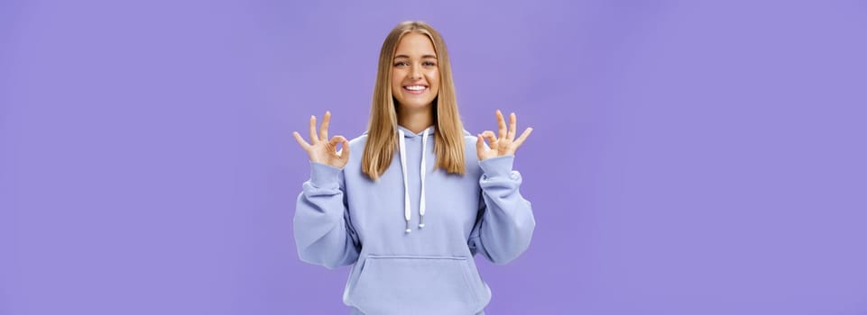 Girl got everything under control. Delighted happy charming woman with fair hair and tanned skin without makeup showing okay gesture smiling assured and pleased posing in hoodie over purple wall.