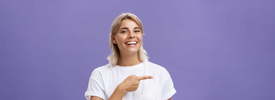 Close-up shot of amused happy and entertained good-looking sociable woman with fair hair and beautiful smile grinning while pointing left with index finger showing awesome copy space over purple wall. Emotions concept
