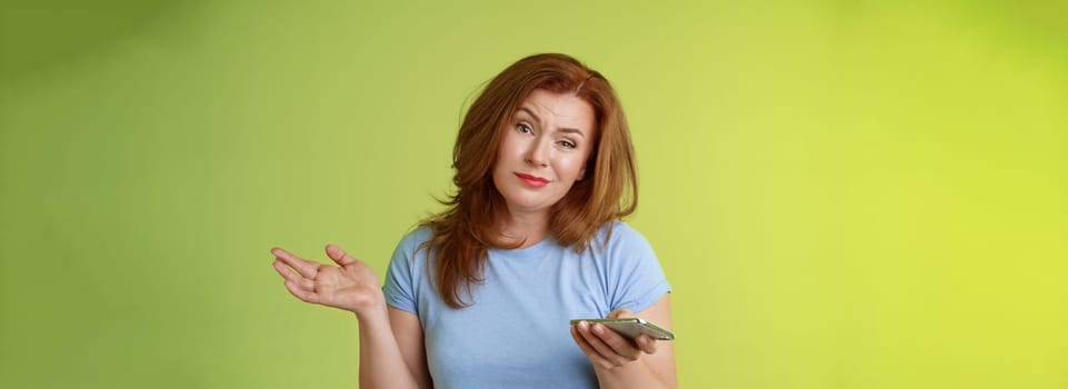 Well meh. Indifferent careless hesitant redhead middle-aged woman mature red female shrugging hold smartphone smirk bored uninterested hold hand aside apathetic attitude green background.