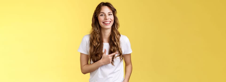 Outgoing charming happy pretty woman curly long hairstyle pointing forefinger left indicate advertisement laughing carefree satisfied hold hand pocket casual friendly pose yellow background.