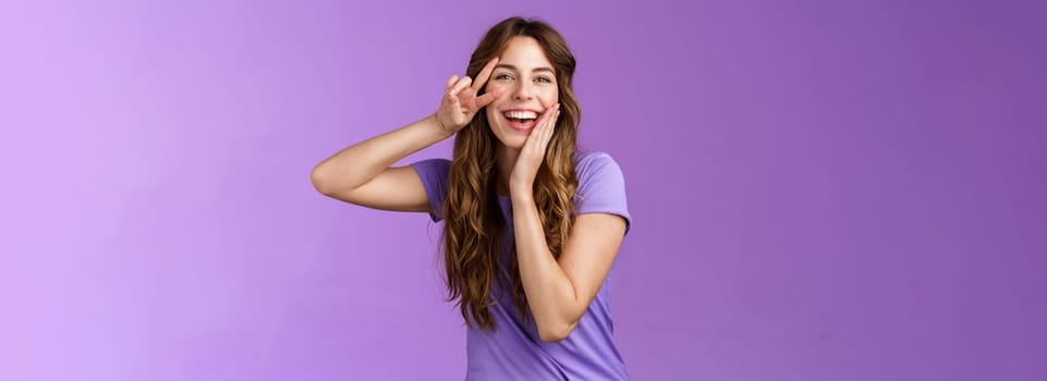 Adorable lovely funny cute woman curly brown hairstyle tilt head touch cheek blishing silly flirty smiling camera show peace victory sign express positive joyful attitude stand purple background. Lifestyle.