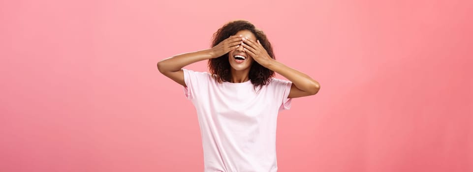 Portrait of charming joyful dark-skinned playful woman with curly hair in t-shirt closing eyes and counting ten with broad happy smile playing hide n seek or waiting for surprise over pink background. Lifestyle.