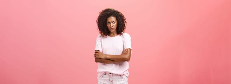 Portrait of offended gloomy sad african american female friend crossing arms on chest in protection gesture frowning looking from under forehead with insult looking envious and angry over pink wall. Lifestyle.