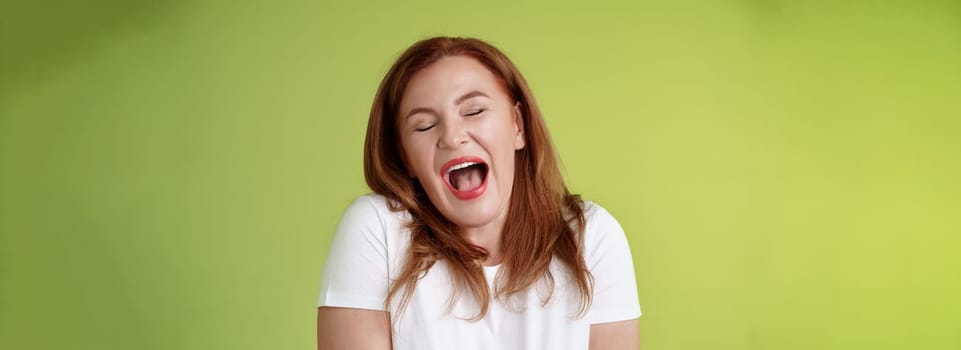 Lazy weekends finally time sleep. Cheerful redhead middle-aged 50s woman yawning satisfied close eyes feel sleepy wake up early morning wanna take nap stand green background watching boring film.