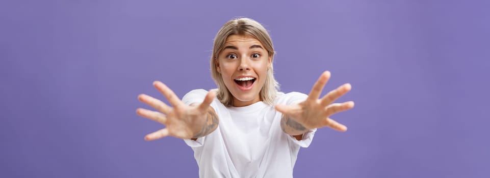 Lifestyle. Waist-up shot of amused and excited attractive stylish young woman in white t-shirt pulling hands at camera with desire smiling thrilled and happy wanting hug or take something over purple background.