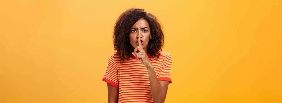 Shh do not disturb. Portrait of serious-looking bossy and displeased cute african american girl with afro hairstyle frowning shushing at camera with index finger over mouth forbidding speak loud.