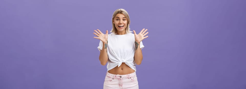 Lifestyle. Indoor shot of delighted and amused attractive stylish urban female with blond hair in stylish white t-shirt and summer shorts raising palms from surprise and amazement standing over purple background.