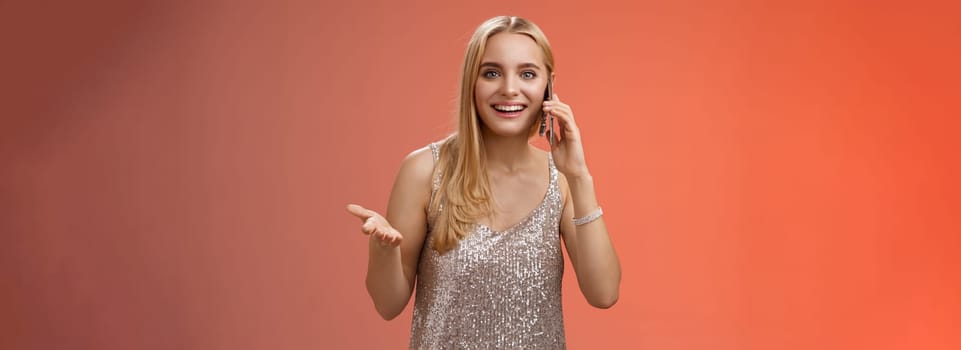 Joyful talkative outgoing attractive blond woman talking friend smartphone gesturing amused smiling broadly retelling fresh rumors after party wearing silver stylish dress, red background.