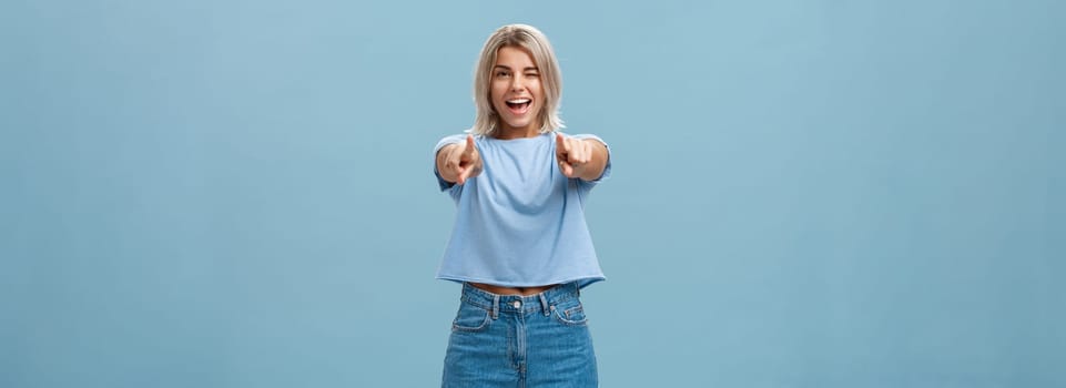 Happy enthusiastic stylish blonde female with tattoos pointing at camera as if picking or making hint smiling joyfully and winking from amazement and happiness posing over blue background. Copy space