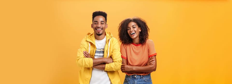 Two african american man and woman being best friends laughing out loud watching funny movie in cinema all dressed up in stylish outfit. standing with hands crossed on chest and amused expression.