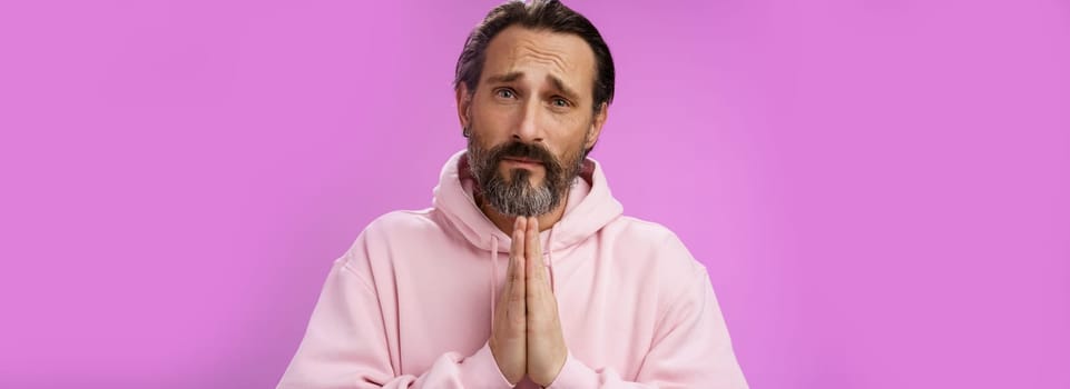Upset handsome bearded mature man need help asking forgiveness supplicating press palms together praying frowning sorrow begging favour lend money, standing purple background miserable.