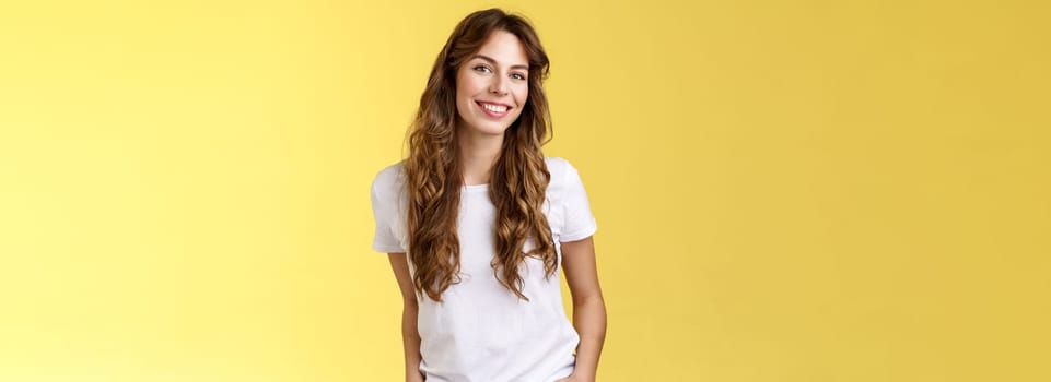 Attractive carefree confident friendly-looking charming feminine ubran woman long curly hair smiling toothy pleased tilt head intrigued hold hands pockets self-assured pose stand yellow background. Lifestyle.