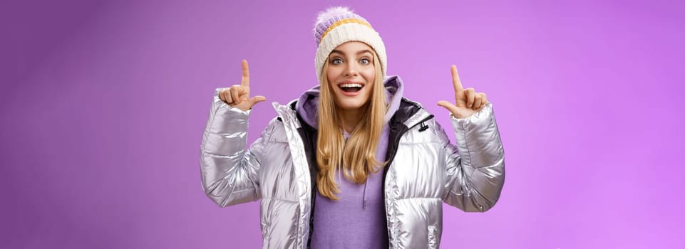 Lifestyle. Excited carefree cheerful fair-haired european girl in silver jacket winter hat raising hands pointing up have excellent idea smiling broadly speaking passionately standing purple background.