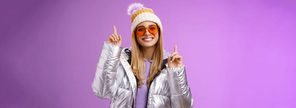 Lifestyle. Joyful energized entertained cute blond woman having fun enjoy vacation snowy mountain trip wearing sunglasses silver jacket winter hat dancing pointing up amused standing purple background.