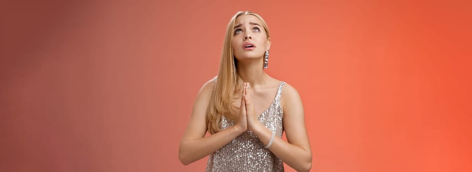 Hopeful worried concerned faithful blond woman in silver dress praying talking god wishing family okay press palms together supplicating nervously begging, standing red background stylish dress.