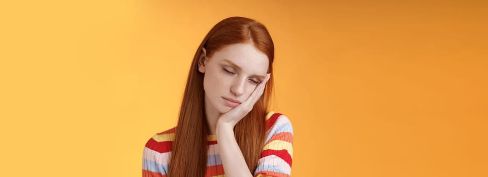 Tired cute redhead female student exhausted feel sleepy fall asleep standing leaning face palm close eyes working part-time night shift, daydreaming lacking energy wanna sleep bed orange background.