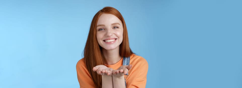 Cute tender kind young ginger girl giving all love you, hold something palms showing camera smiling delighted introduce present grinning romantic gesture send air kisses, blue background.