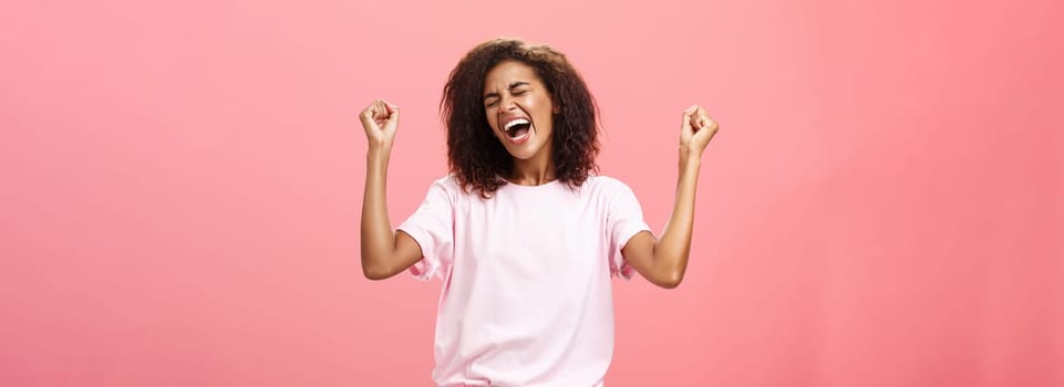 Amazed and happy triumphing african american sportswoman celebrating victory yelling from awesome exciting feelings closing eyes raising fists hight in win gesture standing over pink background. Lifestyle.