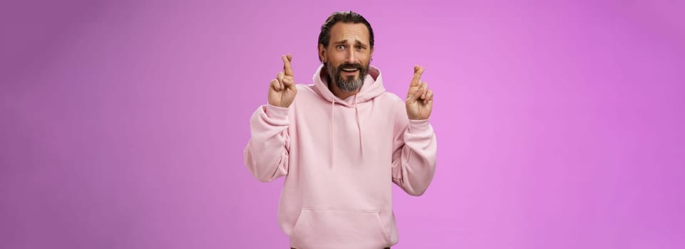 Nervous unsure hopeful handsome bearded 40s man in pink stylish hoodie cringing worried cross figers good luck make wish hope not gonna lose job supplicating praying dream come true. Lifestyle.