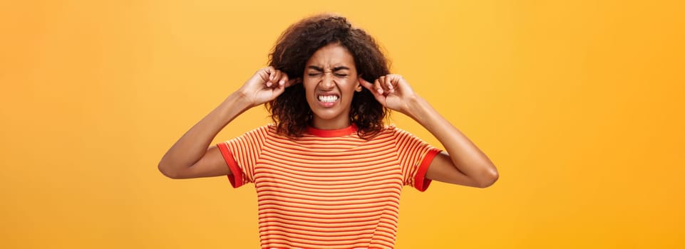 Waist-up shot of intense bothered dark-skinned woman clenchign teeth from discomfort and antipathy closing ears not hear botherind loud noise standing displeased and annoyed over orange wall. Copy space