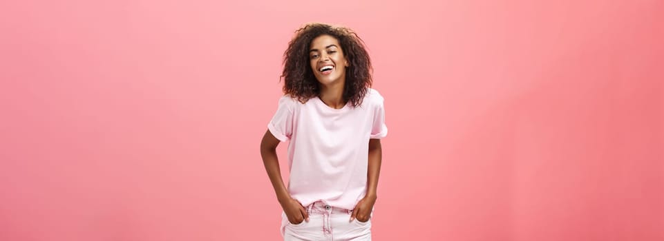 Brighten up life with smile. Carefree charismatic happy african american female with afro hairstyle in trendy outfit laughing out loud from joy and amusement holding hands in pockets over pink wall.