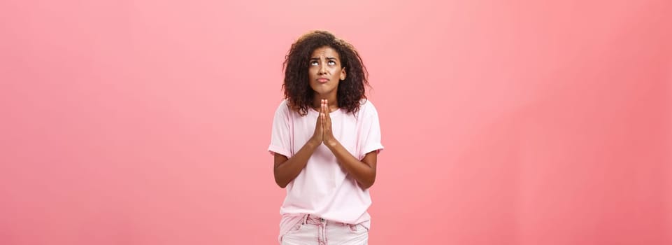 Studio shot of unhappy miserable and hopeless cute african american female holding hands in pray near chest looking up with serious-looking concerned expression making wish to god over pink wall. Lifestyle.