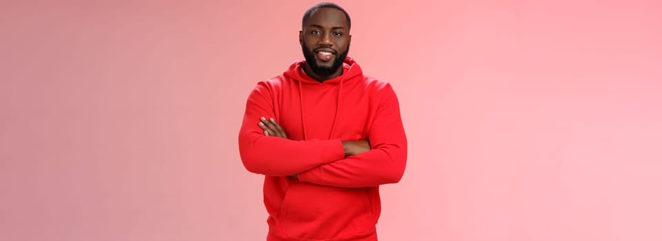 Attractive bearded african-american young guy wearing red hoodie cross arms chest smiling friendly showing friends new place grinning delighted buying own flat, standing proud pink background.