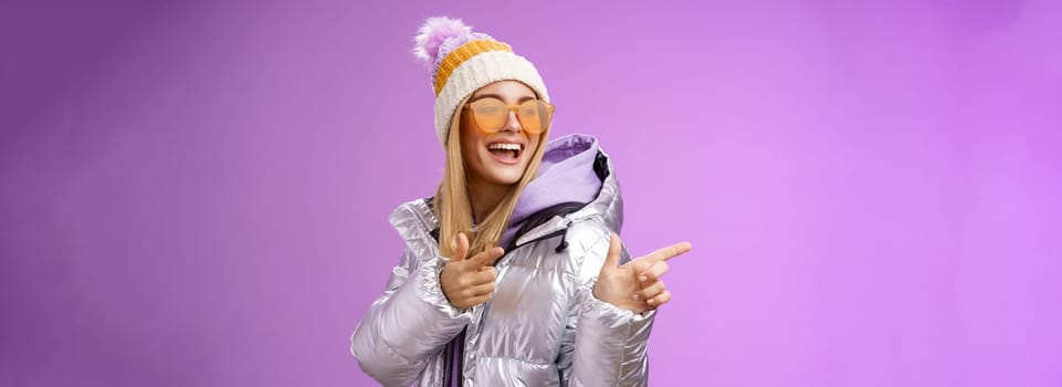 Hey how you doin. Cheeky stylish bond girl having fun greeting friend pointing finger pistols left smiling sassy say hello what up wearing cool silver jacket hat sunglasses, purple background.
