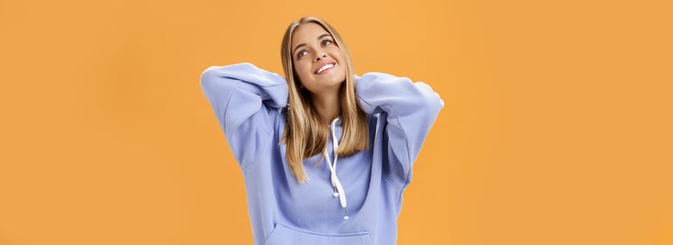 Young pleased and tender woman with tanned skin feeling cozy in warm in trendy hoodie touching back of neck smiling looking at upper right corner dreamy and delighted against orange background. Lifestyle.