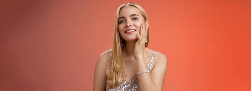 Tender stylish self-assured coquettish young blond wealthy woman in silver shiny dress touching pure clean skin satisfied good makeup get ready party smiling delighted, standing red background.