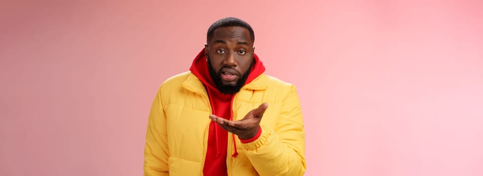 Bothered rude ignorant african-american bearded man pointing palm camera look dumb perplexed, standing confused cannot get clue what happening, wearing yellow jacket, pink background.