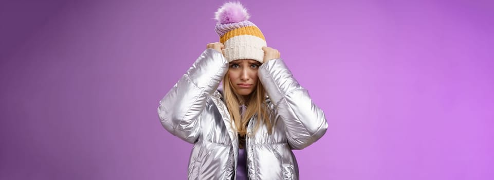 Lifestyle. Upset gloomy insulted cute blond girl complaining offensive behaviour pulling hat forehead frowning sulking sad standing miserable pity purple background in warm stylish glittering silver jacket.