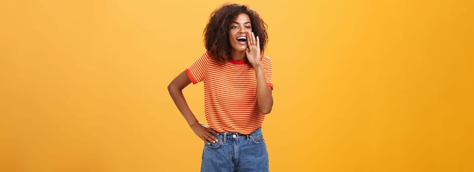 Girl calling sister come to phone yelling out loud holding palm near opened mouth to be scream louder so friend can hear holding arm on waist gazing left with carefree pleased look over orange wall. Lifestyle.