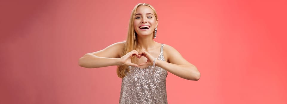 Adorable happy charming lovely blond european girl in silver dress express love positivity show heart gesture confess sympathy passionate feelings towards boyfriend smiling broadly, red background.