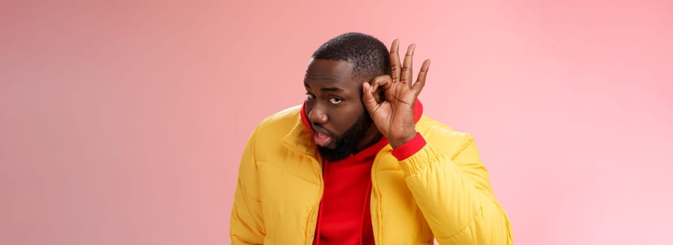 Curious funny charismatic black bearded guy bending forward hold hands ear eavesdropping wanna overhear girlfriend conversation behind door standing curious gossiping pink background.
