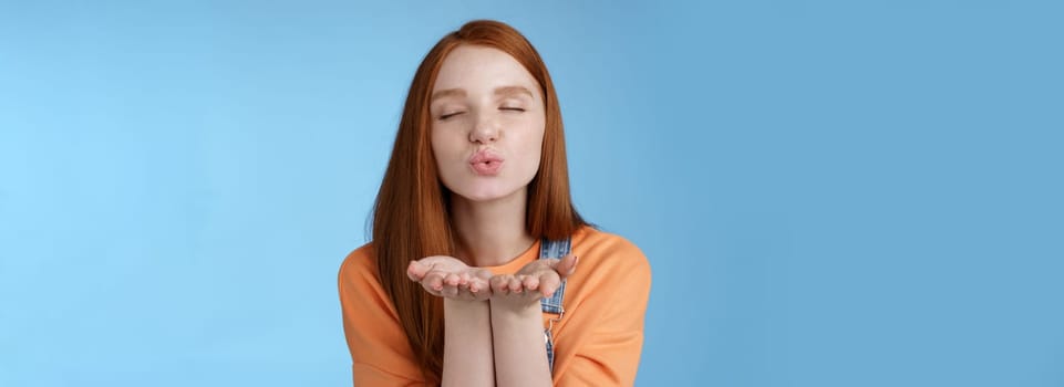Lifestyle. Passionate dreamy pretty redhead girl sending air kisses camera close eyes folding lips hold hands near mouth give muah internet followers recording vlog standing romantic tender blue background.