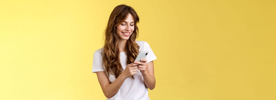 Cheerful lovely girlfriend texting friend pleased cute smile tap smartphone screen smiling broadly look mobile phone display tenderly writing post contemplate touching photo yellow background.