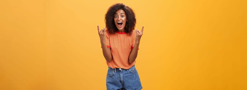 Stylish african american woman being delighted and excited before cool concert showing rock n roll gesture with both hands screaming from satisfaction and happiness cheering over orange background. Copy space