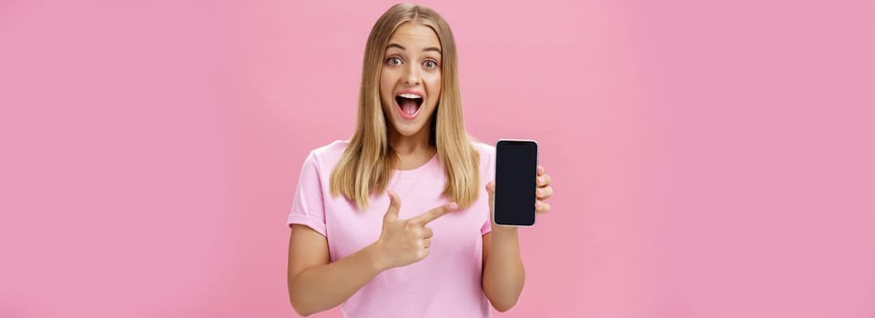 Portrait of excited and amused charming pleased female client using new smartphone advicing friends to buy awesome gadget holding cellphone and pointing at device screen with broad amused smile. Advertisement and technology concept